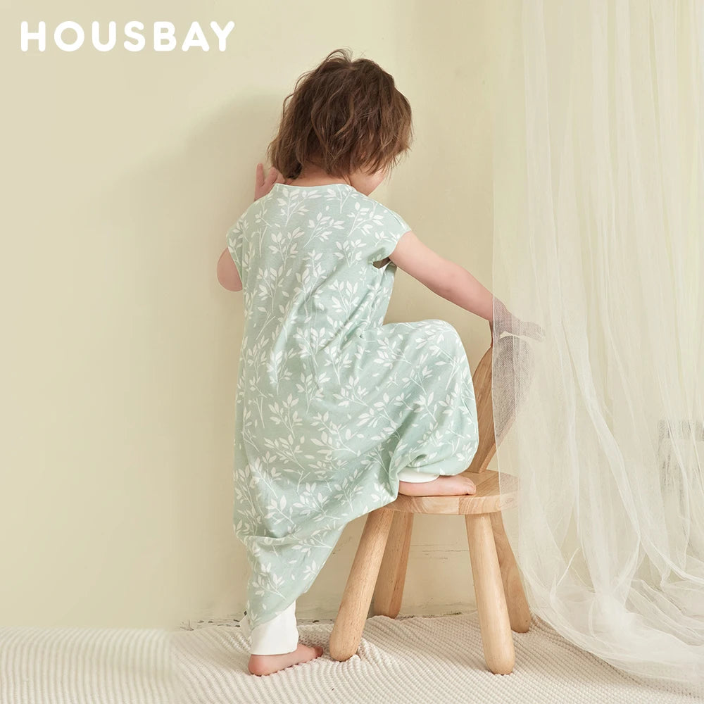 Enfant assis sur un tabouret en bois, portant la gigoteuse à pied vert sauge, photo de marque Housbay